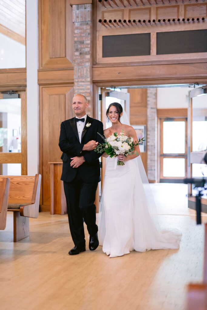 father walk bride down the aisle, arms interlocked, in St. Francis by the Sea Catholic Church during a sea pines wedding