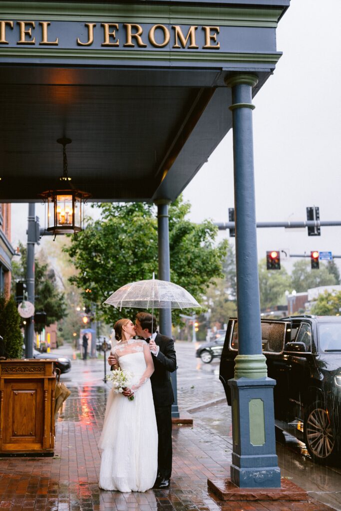 bride and groom kissing under a clear umbrella outside Hotel Jerome during a rainy Aspen wedding