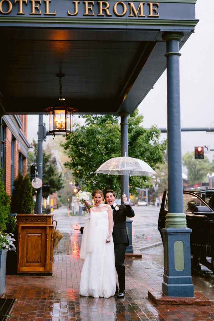 bride and groom celebrating with a clear umbrella in the rain outside Hotel Jerome in Aspen colorado
