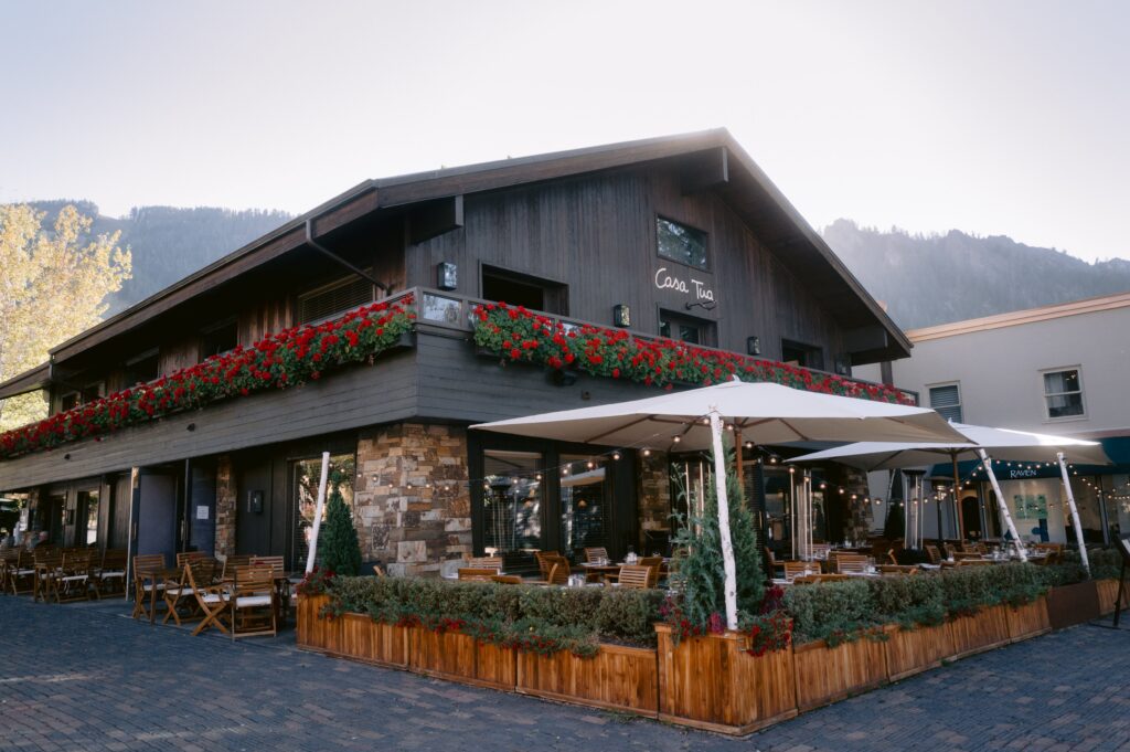 exterior of Casa Tua in Aspen with red geranium-lined balconies, rustic wood and stone façade, and outdoor patio dining beneath white umbrellas at sunset