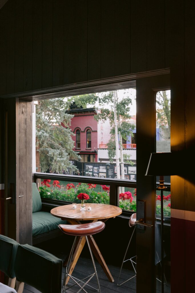 cozy balcony seating at Casa Tua in downtown Aspen featuring a round wooden table, candlelight, red geraniums, and views of historic buildings across the street
