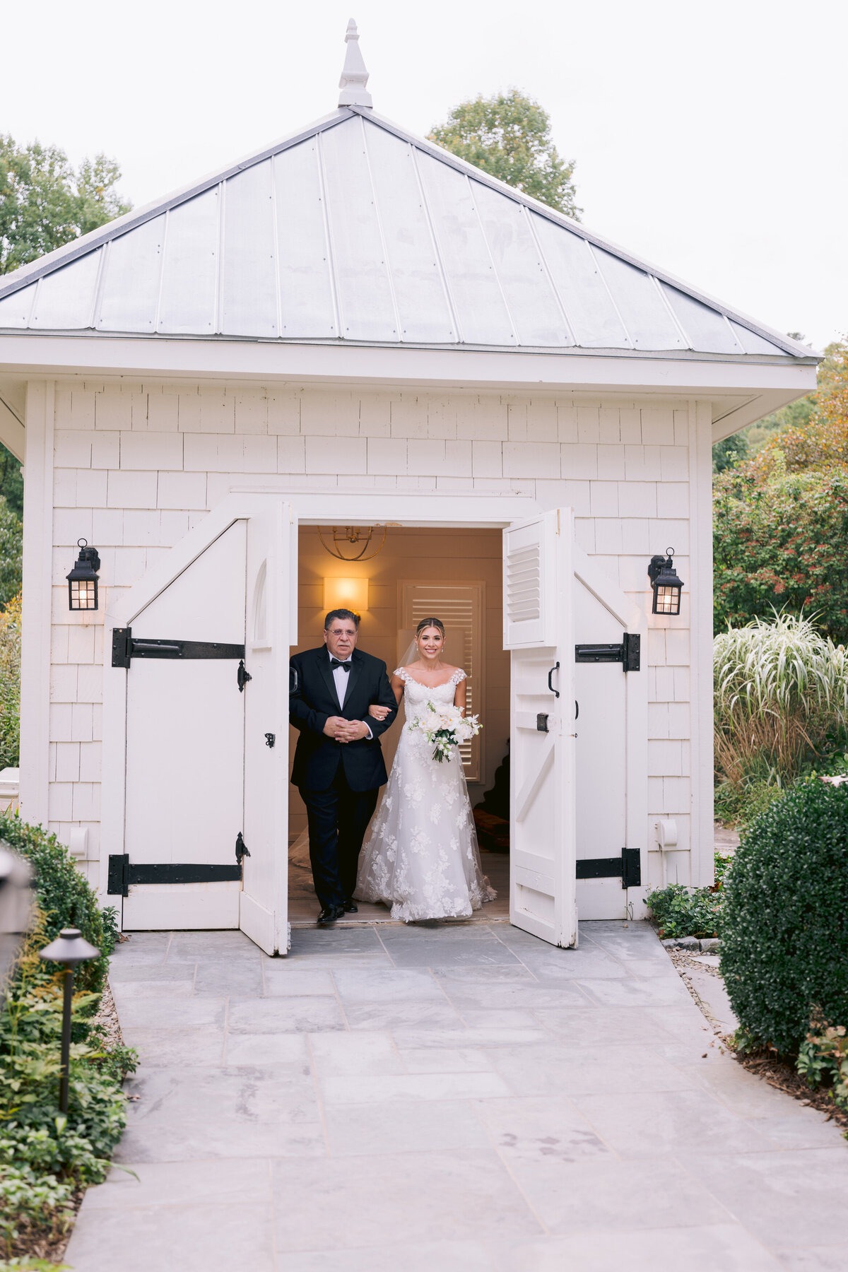 Bride walking arm in arm with her father from a white garden building before the ceremony.