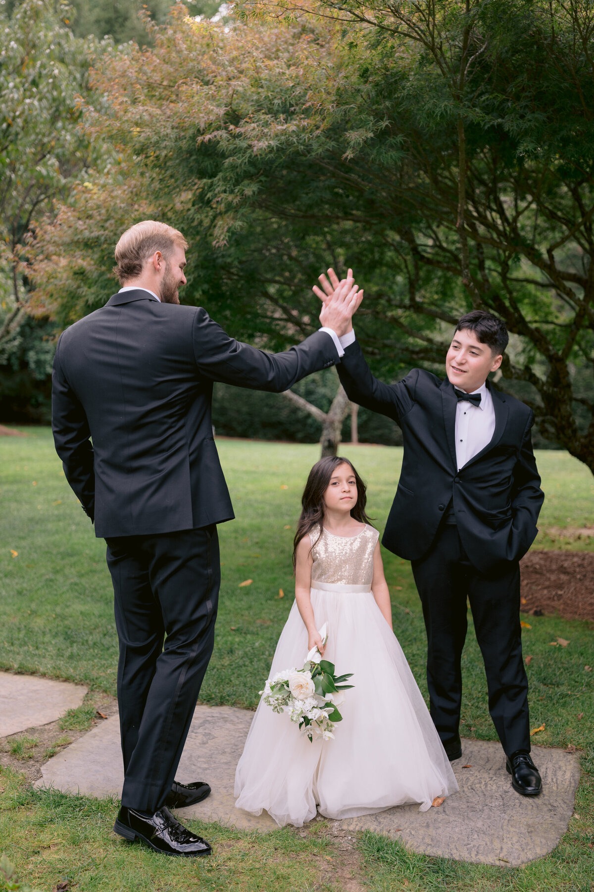 Two groomsmen sharing a high five beside a flower girl holding a white bouquet.