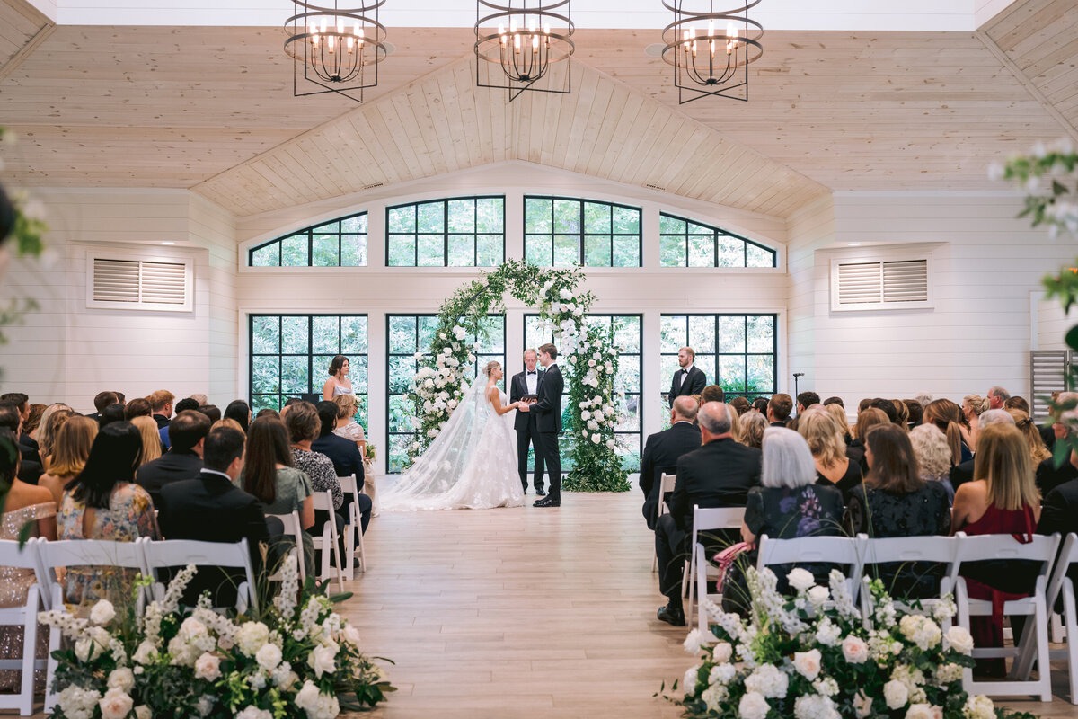 Bride and groom exchanging vows during an indoor Highlands NC wedding ceremony surrounded by guests and white flowers.