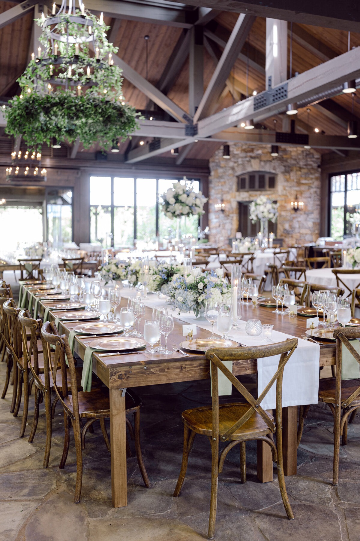 Mountain wedding reception with long wooden tables, white and blue florals, and greenery-covered chandeliers.
